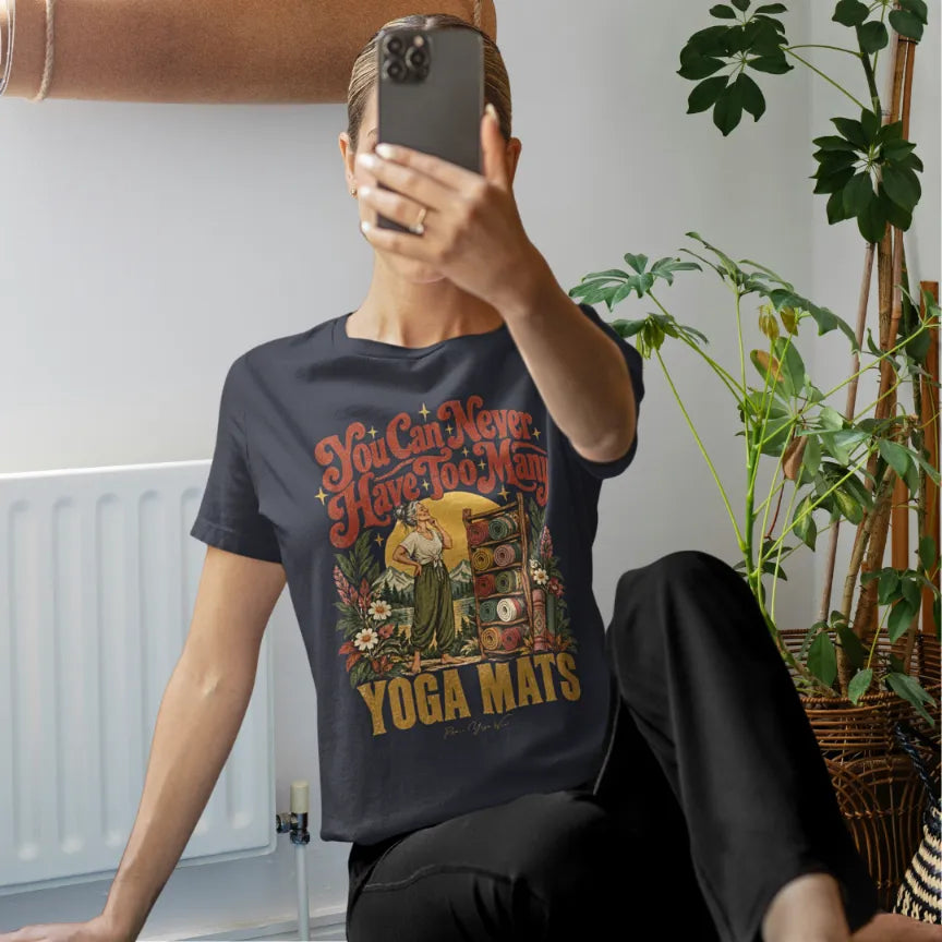 A woman takes a selfie beside indoor plants, on her yoga mat, ready to start class and wearing a Heather Midnight Navy Mats T-Shirt with a retro illustration of a rack of rolled yoga mats and black trousers.
