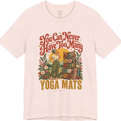 A short sleeved tee called the Mats T-Shirt, flat lay and a soft cotton shirt. Simply says, You Can Never Have Too Many Yoga Mats. Design highlights a silver-haired woman with her hand on her chin, deciding between a wooden rack of rolled yoga mats in rust, mustard, olive, teal and pink, in front of a golden sun, mountains and wildflowers in a retro travel-poster style. Color is Soft Pink.