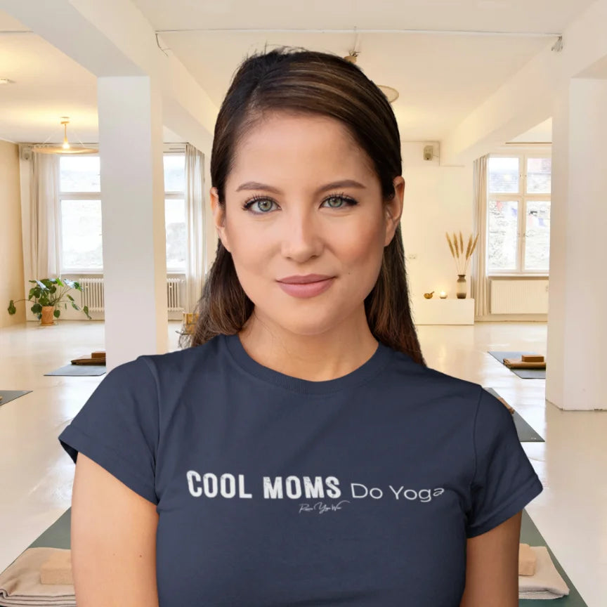 A person with long dark hair pulled back standing in a bright open yoga studio with mats and a plant, wearing the Moms T-Shirt in navy with Cool Moms Do Yoga across the chest.