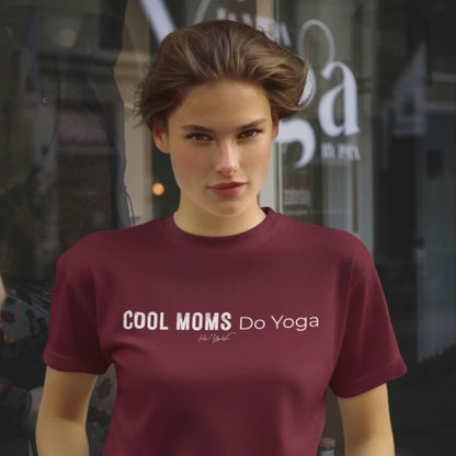 A person with short swept-back light brown hair standing in front of a yoga studio window, wearing the Moms T-Shirt in Heather Cardinal with Cool Moms Do Yoga across the chest.