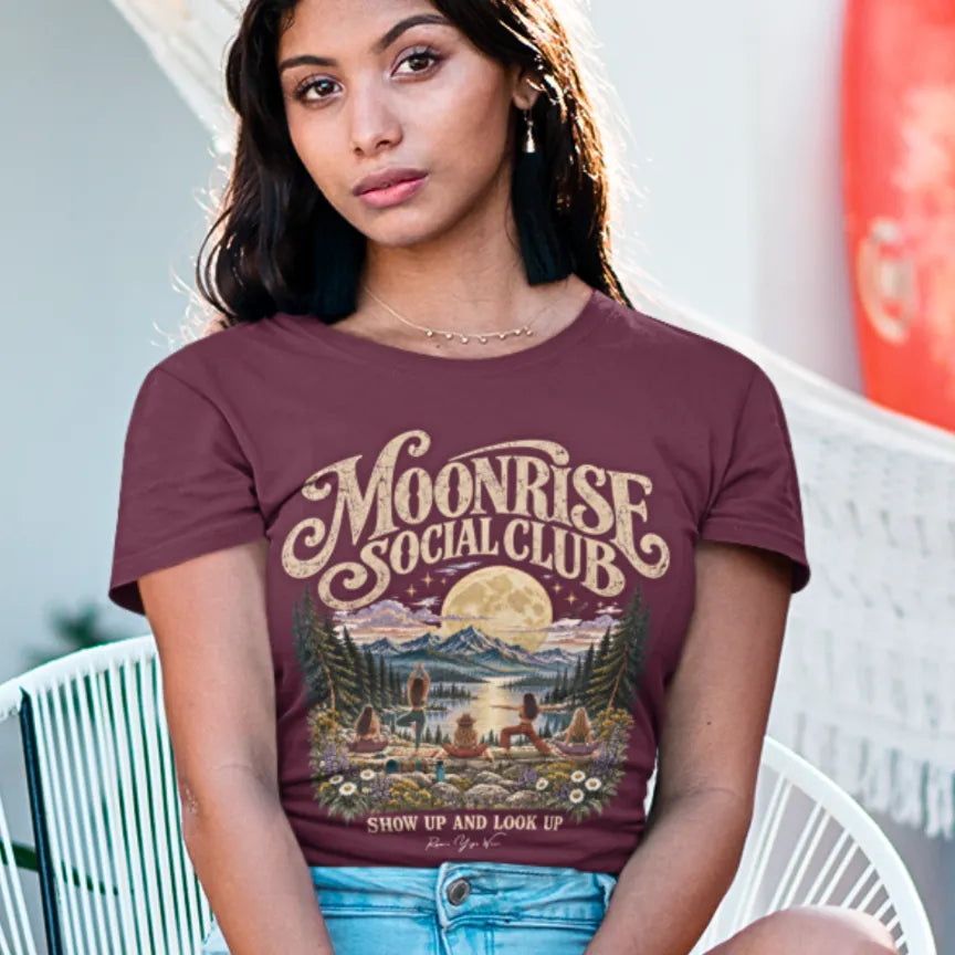 A woman with long dark hair wearing the Moonrise T-Shirt, seated on a white chair outdoors with denim shorts, with the moonlit lake and yoga group design on the front. Color is Heather Cardinal.