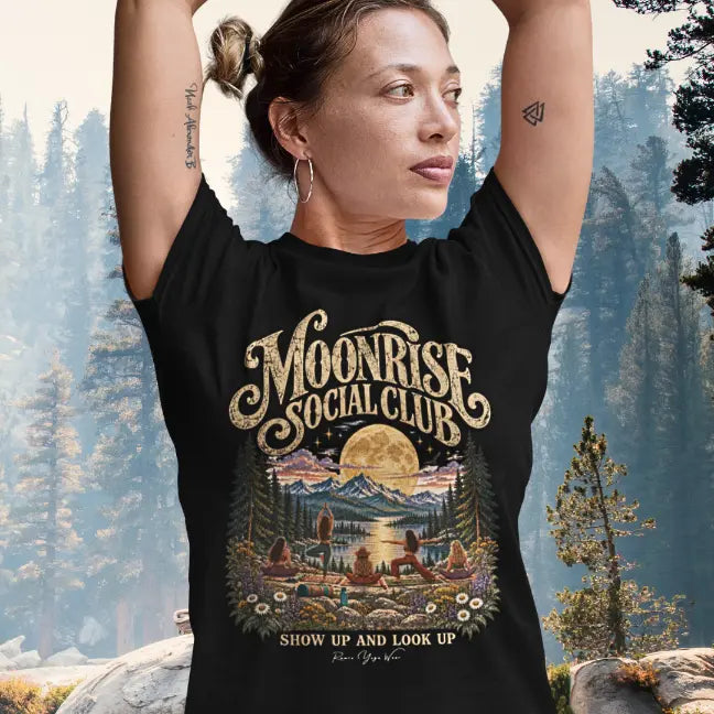 A woman with her hair up and her arms raised overhead wearing the Moonrise T-Shirt, standing in tree pose, in a sunlit pine forest, with the moonlit lake and yoga group design on the front. Color is Black.