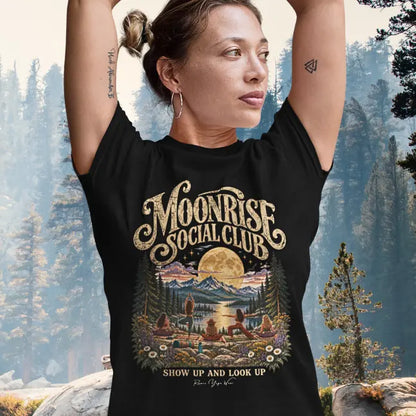A woman with her hair up and her arms raised overhead wearing the Moonrise T-Shirt, standing in tree pose, in a sunlit pine forest, with the moonlit lake and yoga group design on the front. Color is Black.
