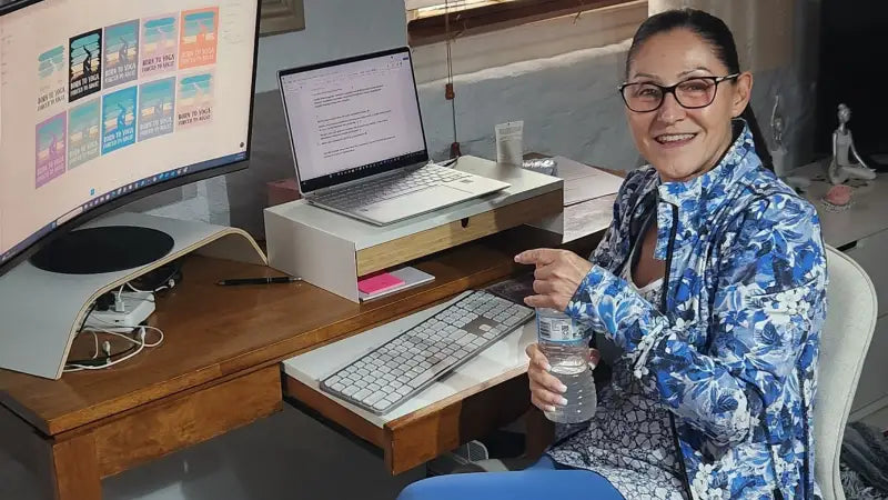 Connie from Ramro Yoga Wear smiling at her desk after yoga practice, holding a water bottle with design mockups open on the computer screen. An authentic, relaxed moment reflecting the brand’s mission to bring intention, comfort, and joy into everyday life.