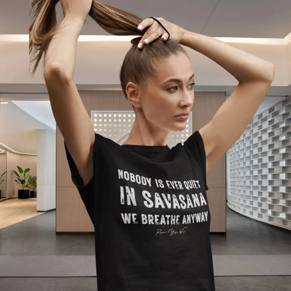 Woman in a black Nobody T-Shirt, hands in her hair, wearing a tee that reads "Nobody is ever quiet in savasana. We breathe anyway." in a bright yoga studio
