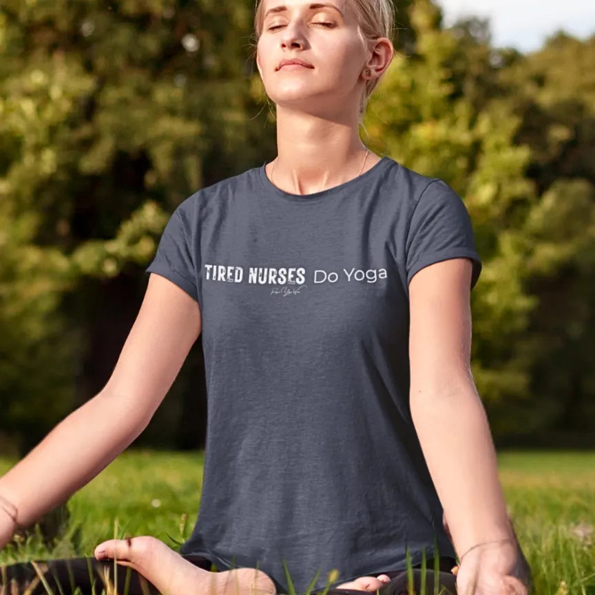 A person sitting cross-legged in a meditation pose on the grass outdoors, eyes closed and face turned to the sun, wearing a Heather Midnight Navy Nurses T-Shirt that says Tired Nurses Do Yoga.