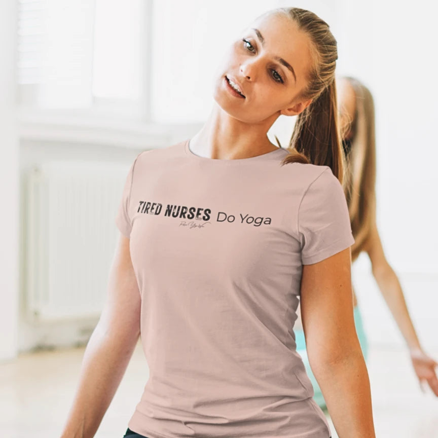 A person with a ponytail stretching and smiling in a bright room, wearing a Heather Prism Peach Nurses T-Shirt that says Tired Nurses Do Yoga.