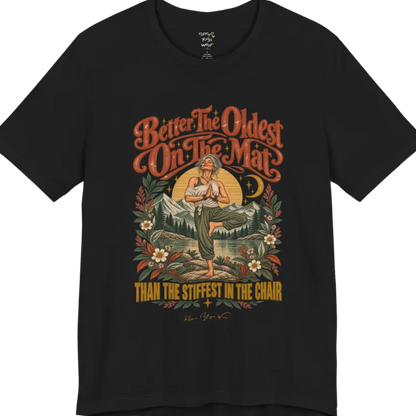 A short sleeved teed has an Illustration of an older woman in tree pose with hands in prayer position, standing on a rock by a lake, framed by mountains, pine trees, a sunburst, and botanical flowers. Oldest T-Shirt flat lay is a Bella+Canvas 3001 and the color is Black.