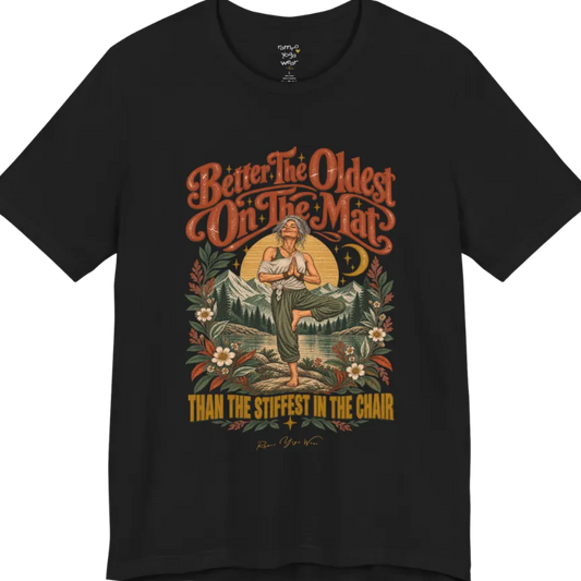 A short sleeved teed has an Illustration of an older woman in tree pose with hands in prayer position, standing on a rock by a lake, framed by mountains, pine trees, a sunburst, and botanical flowers. Oldest T-Shirt flat lay is a Bella+Canvas 3001 and the color is Black.