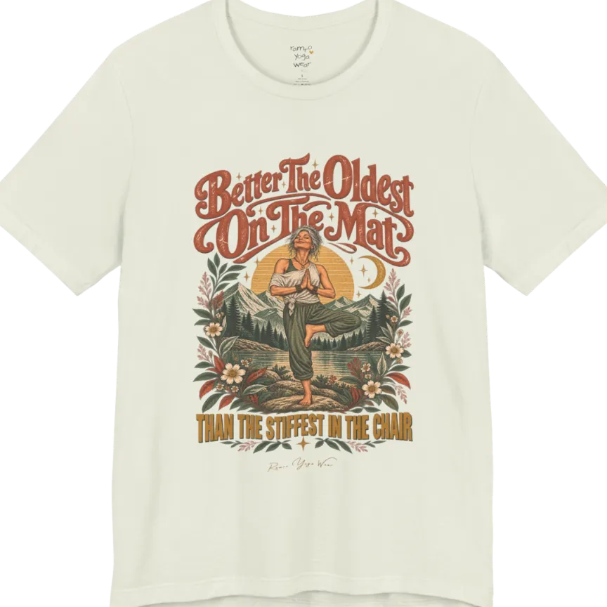 A short sleeved teed has an Illustration of an older woman in tree pose with hands in prayer position, standing on a rock by a lake, framed by mountains, pine trees, a sunburst, and botanical flowers. Oldest T-Shirt flat lay is a Bella+Canvas 3001 and the color is Citron.