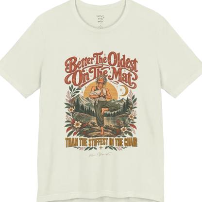 A short sleeved teed has an Illustration of an older woman in tree pose with hands in prayer position, standing on a rock by a lake, framed by mountains, pine trees, a sunburst, and botanical flowers. Oldest T-Shirt flat lay is a Bella+Canvas 3001 and the color is Citron.