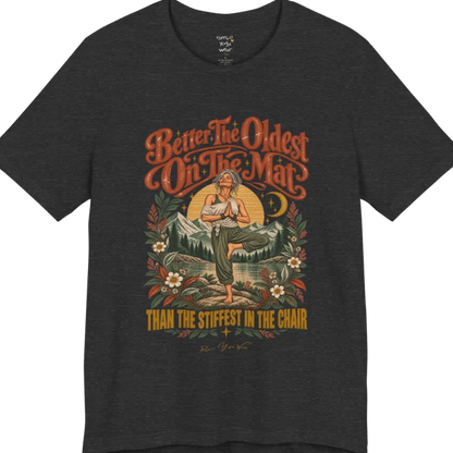 A short sleeved teed has an Illustration of an older woman in tree pose with hands in prayer position, standing on a rock by a lake, framed by mountains, pine trees, a sunburst, and botanical flowers. Oldest T-Shirt flat lay is a Bella+Canvas 3001 and the color is Dark Grey Heather.