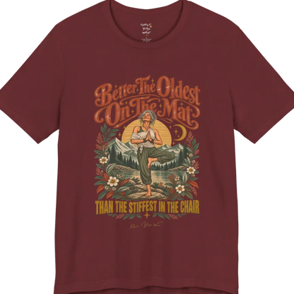 A short sleeved teed has an Illustration of an older woman in tree pose with hands in prayer position, standing on a rock by a lake, framed by mountains, pine trees, a sunburst, and botanical flowers. Oldest T-Shirt flat lay is a Bella+Canvas 3001 and the color is Heather Cardinal.