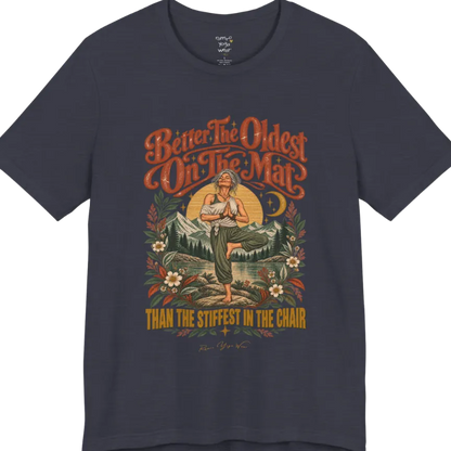 A short sleeved teed has an Illustration of an older woman in tree pose with hands in prayer position, standing on a rock by a lake, framed by mountains, pine trees, a sunburst, and botanical flowers. Oldest T-Shirt flat lay is a Bella+Canvas 3001 and the color is Heather Midnight Navy.