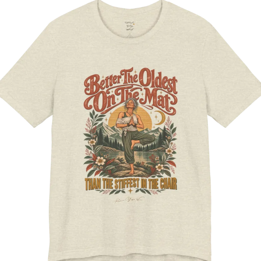 A short sleeved teed has an Illustration of an older woman in tree pose with hands in prayer position, standing on a rock by a lake, framed by mountains, pine trees, a sunburst, and botanical flowers. Oldest T-Shirt flat lay is a Bella+Canvas 3001 and the color is Heather Prism Natural.