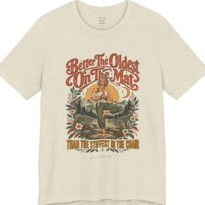 A short sleeved teed has an Illustration of an older woman in tree pose with hands in prayer position, standing on a rock by a lake, framed by mountains, pine trees, a sunburst, and botanical flowers. Oldest T-Shirt flat lay is a Bella+Canvas 3001 and the color is Heather Prism Natural.