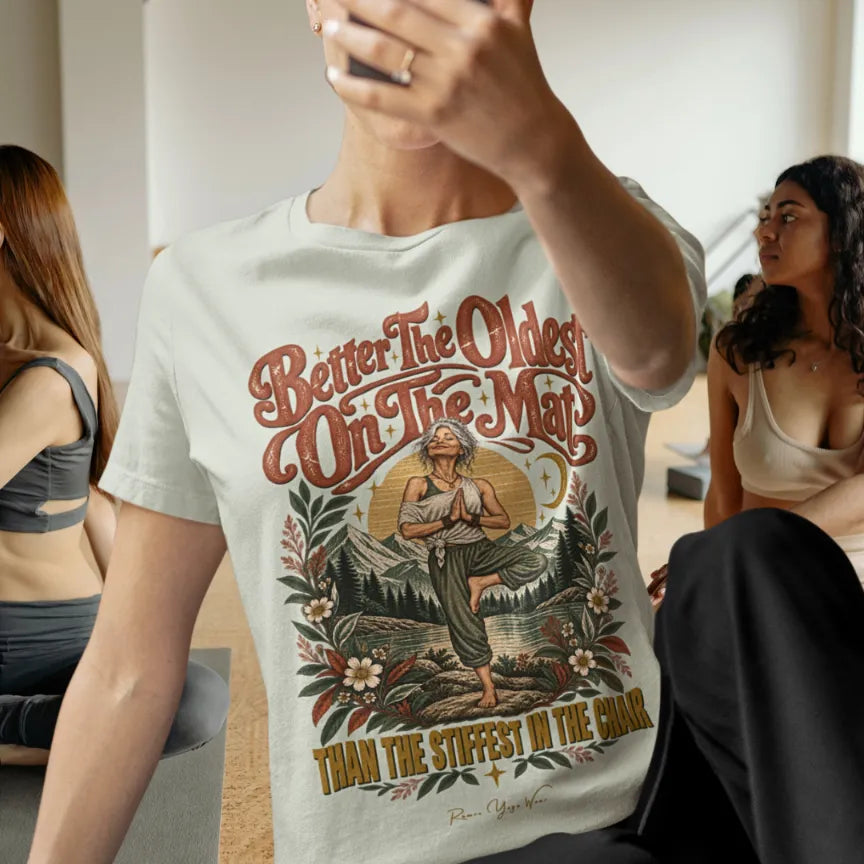 A person in a bright yoga studio is taking a selfie while others are practicing nearby, wearing the Oldest T-Shirt. Oldest T-Shirt is a Bella+Canvas 3001 and the color is Citron.