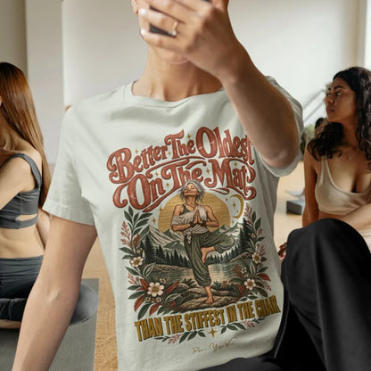 A person in a bright yoga studio is taking a selfie while others are practicing nearby, wearing the Oldest T-Shirt. Oldest T-Shirt is a Bella+Canvas 3001 and the color is Citron.