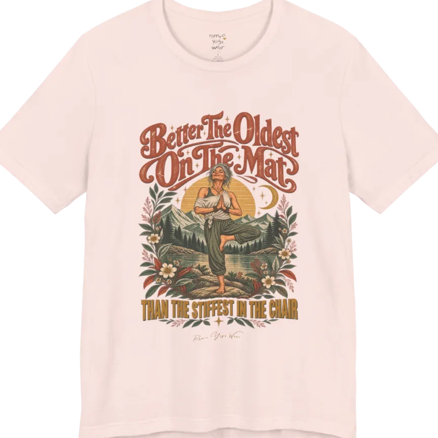 A short sleeved teed has an Illustration of an older woman in tree pose with hands in prayer position, standing on a rock by a lake, framed by mountains, pine trees, a sunburst, and botanical flowers. Oldest T-Shirt flat lay is a Bella+Canvas 3001 and the color is Soft Pink.