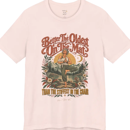 A short sleeved teed has an Illustration of an older woman in tree pose with hands in prayer position, standing on a rock by a lake, framed by mountains, pine trees, a sunburst, and botanical flowers. Oldest T-Shirt flat lay is a Bella+Canvas 3001 and the color is Soft Pink.