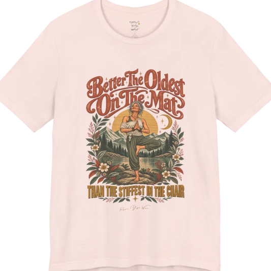 A short sleeved teed has an Illustration of an older woman in tree pose with hands in prayer position, standing on a rock by a lake, framed by mountains, pine trees, a sunburst, and botanical flowers. Oldest T-Shirt flat lay is a Bella+Canvas 3001 and the color is Soft Pink.