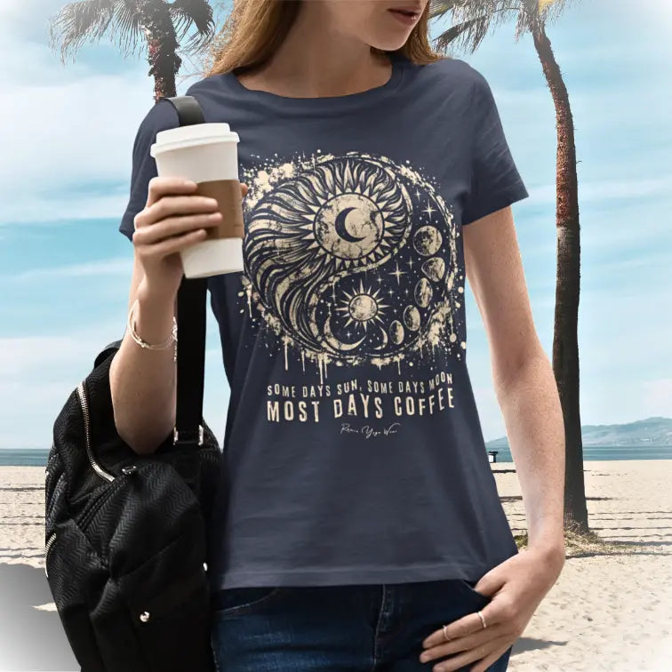 A person with light brown hair holding a takeaway coffee cup and carrying a black bag on a sunny beach with palm trees, wearing the Phases T-Shirt. The tee shows a grunge sun and moon yin yang in cream with the words Some Days Sun, Some Days Moon, Most Days Coffee. Color is heather midnight navy.