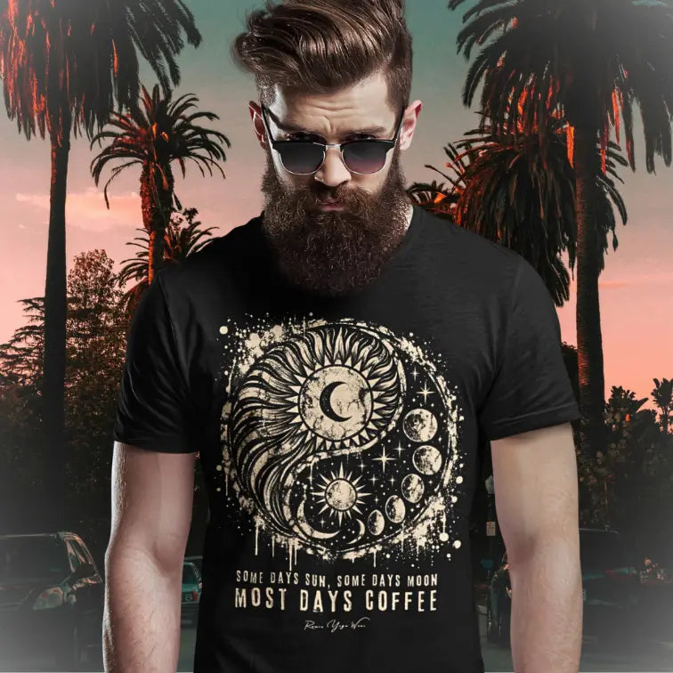A bearded person in sunglasses with styled hair standing among palm trees at sunset on a Californian street, wearing the Phases T-Shirt. The tee shows a grunge sun and moon yin yang in cream with the words Some Days Sun, Some Days Moon, Most Days Coffee. Color is black.