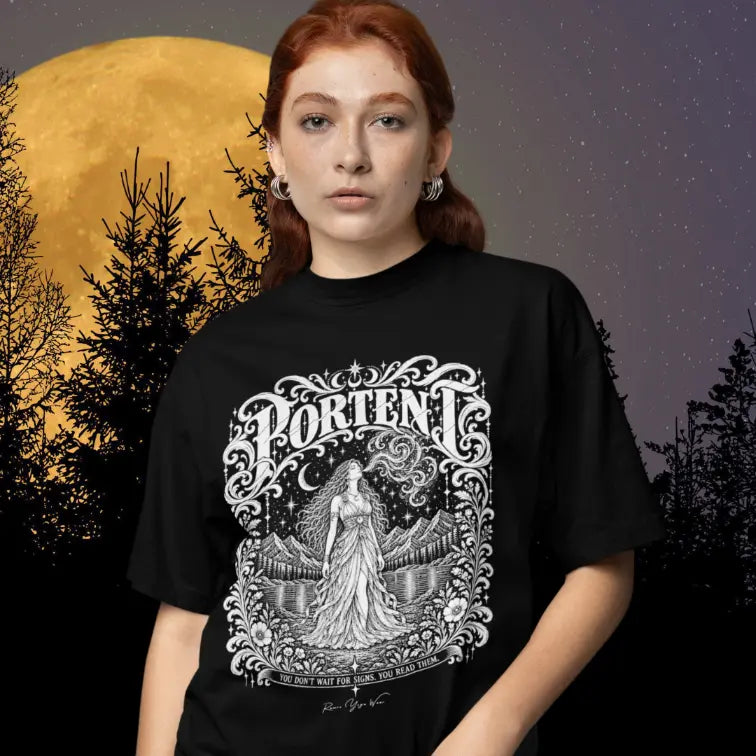 A person with auburn hair and silver hoop earrings, wearing a black Portent T-Shirt with a white engraved figure and crescent moon design, standing against a golden full moon and a silhouette of pine trees.