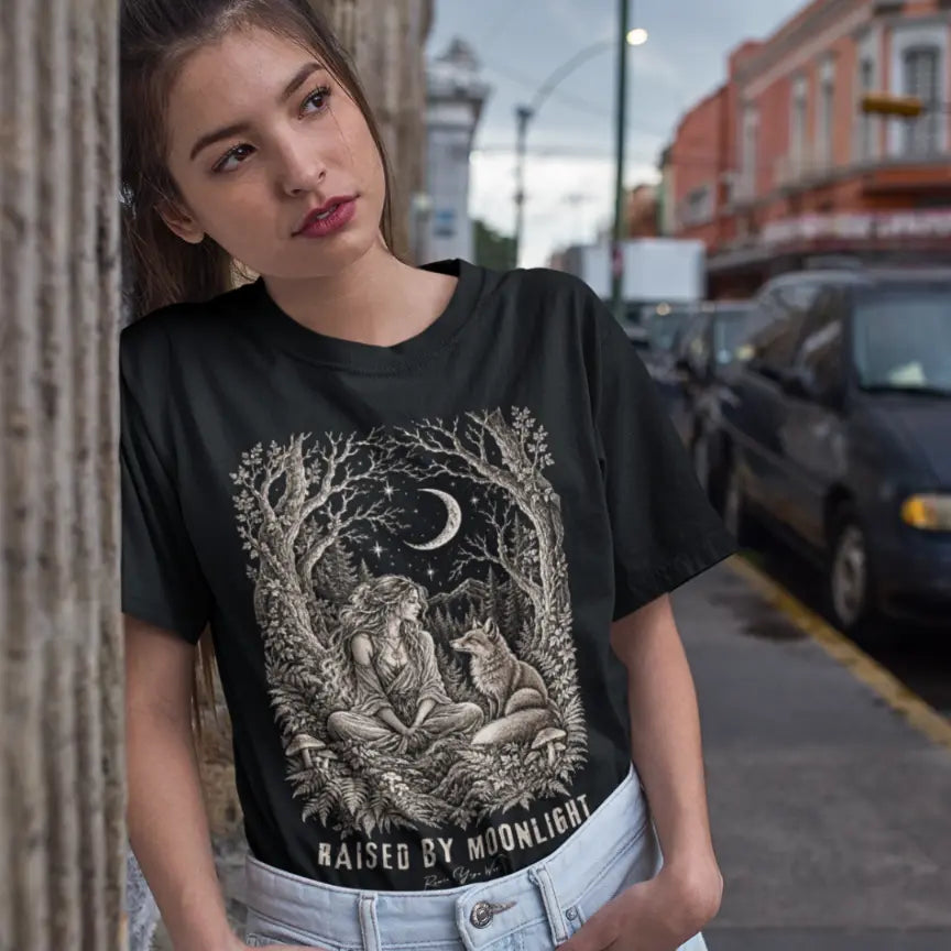 A short sleeved tee called the Raised T-Shirt, Gildan 64000 shirt, worn on a model leaning against a stone wall on a city street, paired with blue jeans. Simply says, Raised By Moonlight. Design highlights a woman sitting cross-legged in a moonlit forest clearing with a fox beside her, surrounded by mushrooms and ferns, in fine sepia linework. Color is Black.