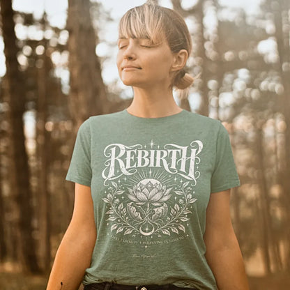 A woman with her eyes closed stands among sunlit pine trees, wearing the Rebirth T-Shirt. Rebirth T-Shirt is a Gildan 64000 and the color is Sage.