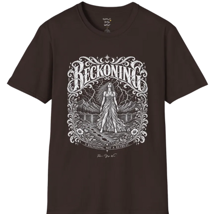 A short sleeved tee called the Reckoning T-Shirt, flat lay and a soft cotton shirt. Simply says, A reckoning, not a retreat. Design highlights ornate white RECKONING lettering framed by flourishes and wild florals, with a long-haired figure in a flowing gown walking a footprint path toward a lightning storm over mountains and a lake. Color is Dark Chocolate.