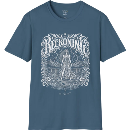 A short sleeved tee called the Reckoning T-Shirt, flat lay and a soft cotton shirt. Simply says, A reckoning, not a retreat. Design highlights ornate white RECKONING lettering framed by flourishes and wild florals, with a long-haired figure in a flowing gown walking a footprint path toward a lightning storm over mountains and a lake. Color is Indigo Blue.
