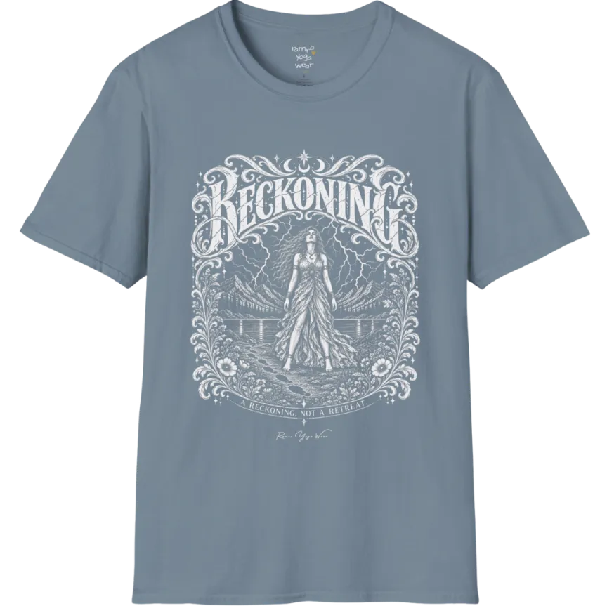 A short sleeved tee called the Reckoning T-Shirt, flat lay and a soft cotton shirt. Simply says, A reckoning, not a retreat. Design highlights ornate white RECKONING lettering framed by flourishes and wild florals, with a long-haired figure in a flowing gown walking a footprint path toward a lightning storm over mountains and a lake. Color is Stone Blue.