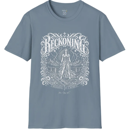 A short sleeved tee called the Reckoning T-Shirt, flat lay and a soft cotton shirt. Simply says, A reckoning, not a retreat. Design highlights ornate white RECKONING lettering framed by flourishes and wild florals, with a long-haired figure in a flowing gown walking a footprint path toward a lightning storm over mountains and a lake. Color is Stone Blue.