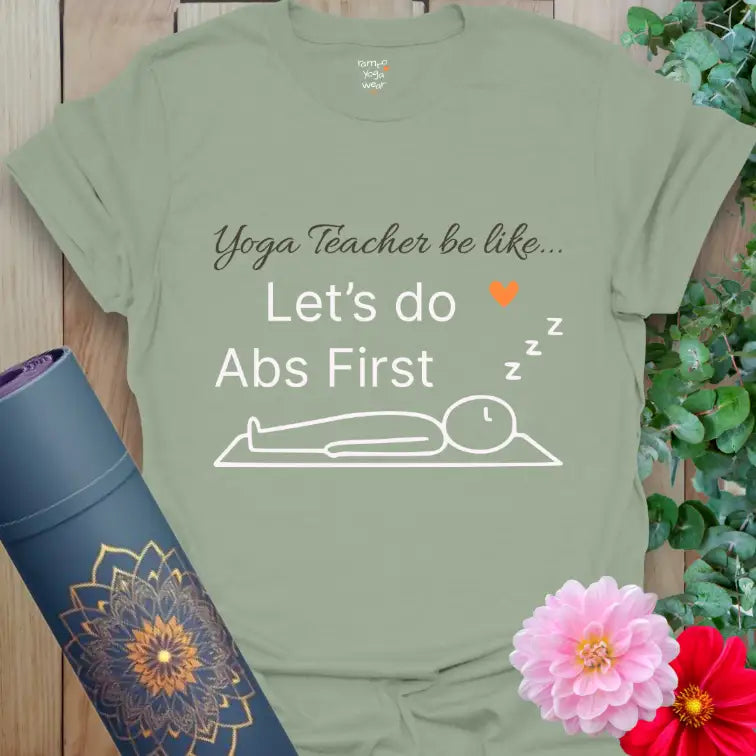 Sage Abs First T-Shirt with playful yoga quote “Yoga Teacher be like… Let’s do Abs First” and minimalist line-art figure resting in Savasana. Calming green tone with white text and Ramro orange heart accent.