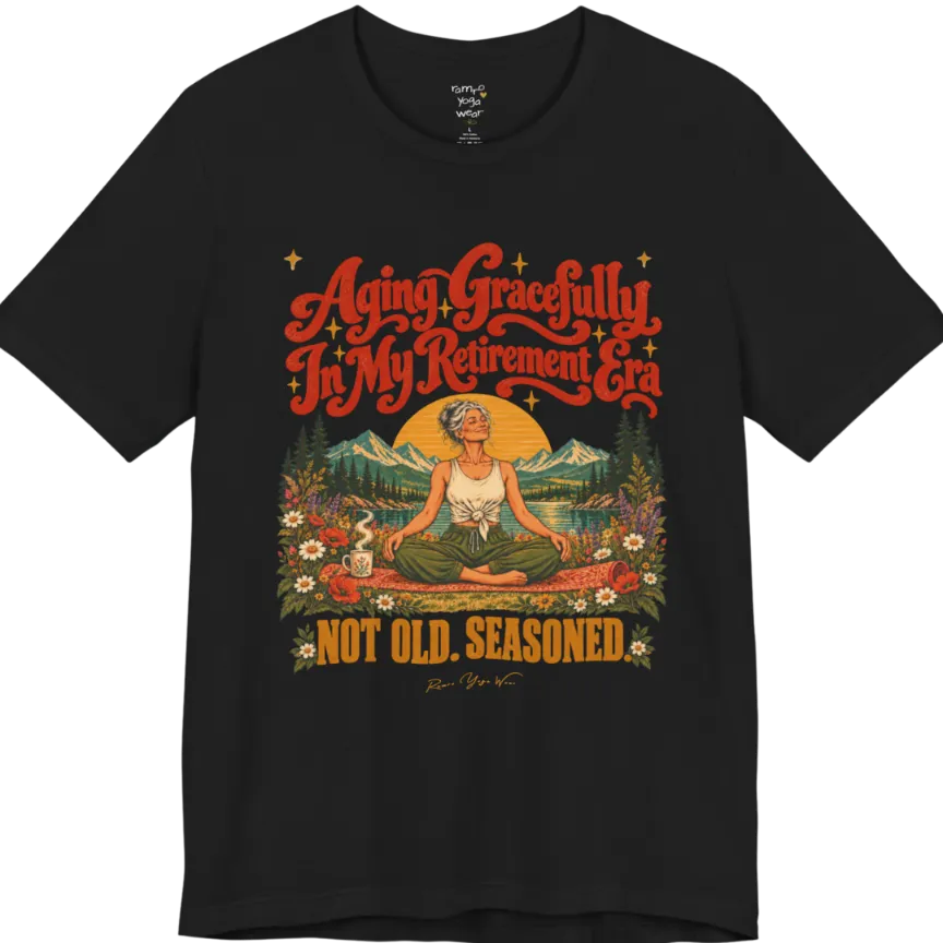 A short sleeved tee called the Seasoned T-Shirt, flat lay and a soft cotton blend shirt. Simply says, Aging Gracefully In My Retirement Era. Not Old. Seasoned. Design highlights a silver-haired figure sitting cross-legged in easy pose on a patterned mat beside a steaming mug, with a rising sun, mountains, a lake and wildflowers, and gold sparkle stars around the script lettering. Color is Black.