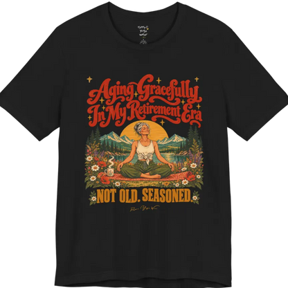 A short sleeved tee called the Seasoned T-Shirt, flat lay and a soft cotton blend shirt. Simply says, Aging Gracefully In My Retirement Era. Not Old. Seasoned. Design highlights a silver-haired figure sitting cross-legged in easy pose on a patterned mat beside a steaming mug, with a rising sun, mountains, a lake and wildflowers, and gold sparkle stars around the script lettering. Color is Black.