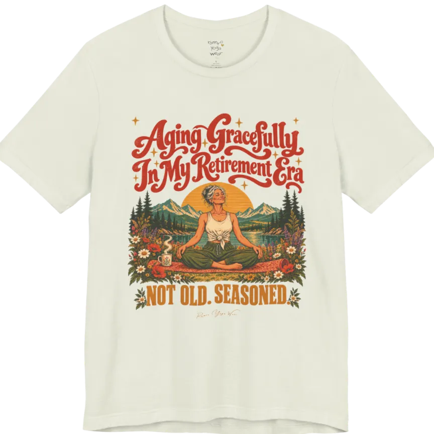 A short sleeved tee called the Seasoned T-Shirt, flat lay and a soft cotton blend shirt. Simply says, Aging Gracefully In My Retirement Era. Not Old. Seasoned. Design highlights a silver-haired figure sitting cross-legged in easy pose on a patterned mat beside a steaming mug, with a rising sun, mountains, a lake and wildflowers, and gold sparkle stars around the script lettering. Color is Citron.