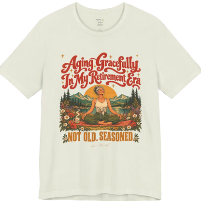A short sleeved tee called the Seasoned T-Shirt, flat lay and a soft cotton blend shirt. Simply says, Aging Gracefully In My Retirement Era. Not Old. Seasoned. Design highlights a silver-haired figure sitting cross-legged in easy pose on a patterned mat beside a steaming mug, with a rising sun, mountains, a lake and wildflowers, and gold sparkle stars around the script lettering. Color is Citron.