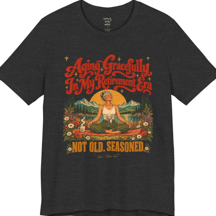 A short sleeved tee called the Seasoned T-Shirt, flat lay and a soft cotton blend shirt. Simply says, Aging Gracefully In My Retirement Era. Not Old. Seasoned. Design highlights a silver-haired figure sitting cross-legged in easy pose on a patterned mat beside a steaming mug, with a rising sun, mountains, a lake and wildflowers, and gold sparkle stars around the script lettering. Color is Dark Grey Heather.