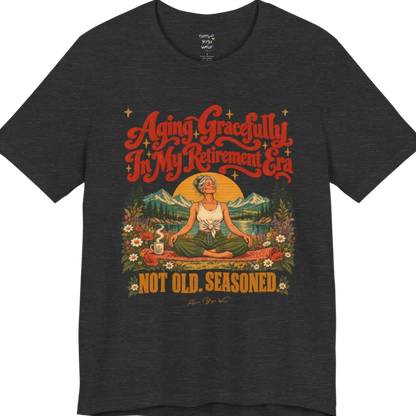 A short sleeved tee called the Seasoned T-Shirt, flat lay and a soft cotton blend shirt. Simply says, Aging Gracefully In My Retirement Era. Not Old. Seasoned. Design highlights a silver-haired figure sitting cross-legged in easy pose on a patterned mat beside a steaming mug, with a rising sun, mountains, a lake and wildflowers, and gold sparkle stars around the script lettering. Color is Dark Grey Heather.