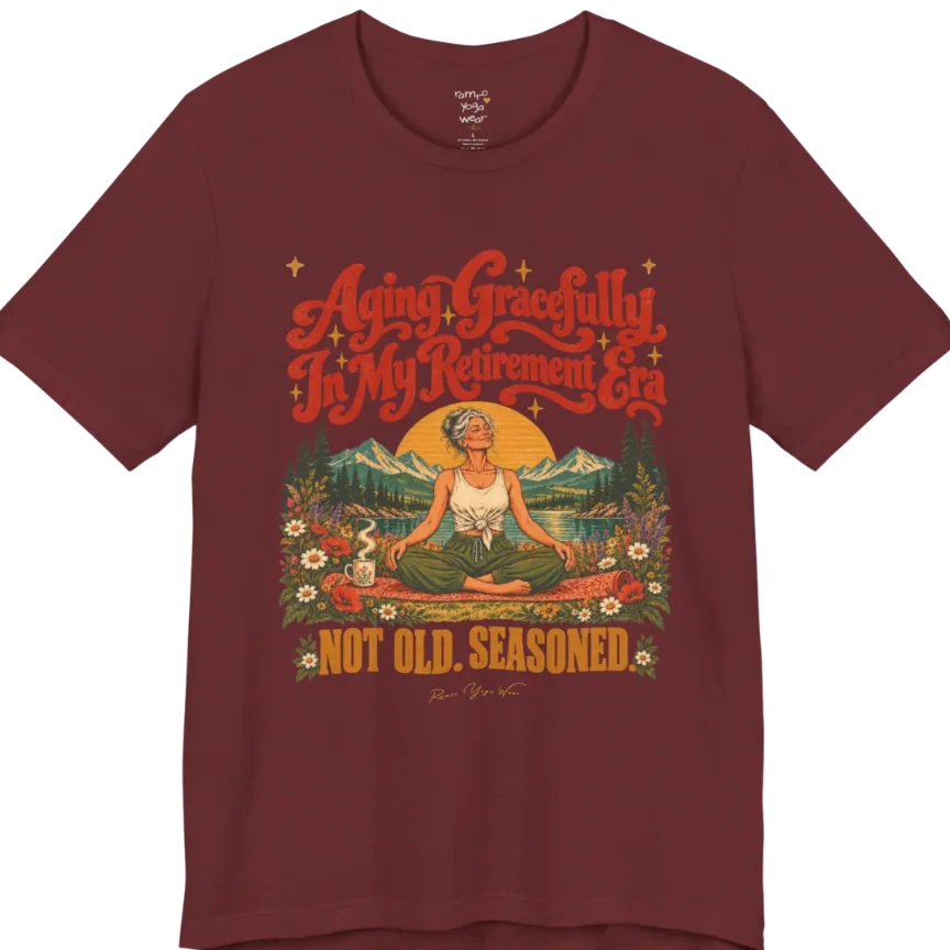 A short sleeved tee called the Seasoned T-Shirt, flat lay and a soft cotton blend shirt. Simply says, Aging Gracefully In My Retirement Era. Not Old. Seasoned. Design highlights a silver-haired figure sitting cross-legged in easy pose on a patterned mat beside a steaming mug, with a rising sun, mountains, a lake and wildflowers, and gold sparkle stars around the script lettering. Color is Heather Cardinal.