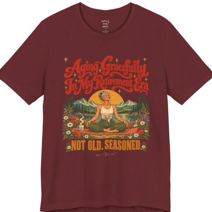 A short sleeved tee called the Seasoned T-Shirt, flat lay and a soft cotton blend shirt. Simply says, Aging Gracefully In My Retirement Era. Not Old. Seasoned. Design highlights a silver-haired figure sitting cross-legged in easy pose on a patterned mat beside a steaming mug, with a rising sun, mountains, a lake and wildflowers, and gold sparkle stars around the script lettering. Color is Heather Cardinal.