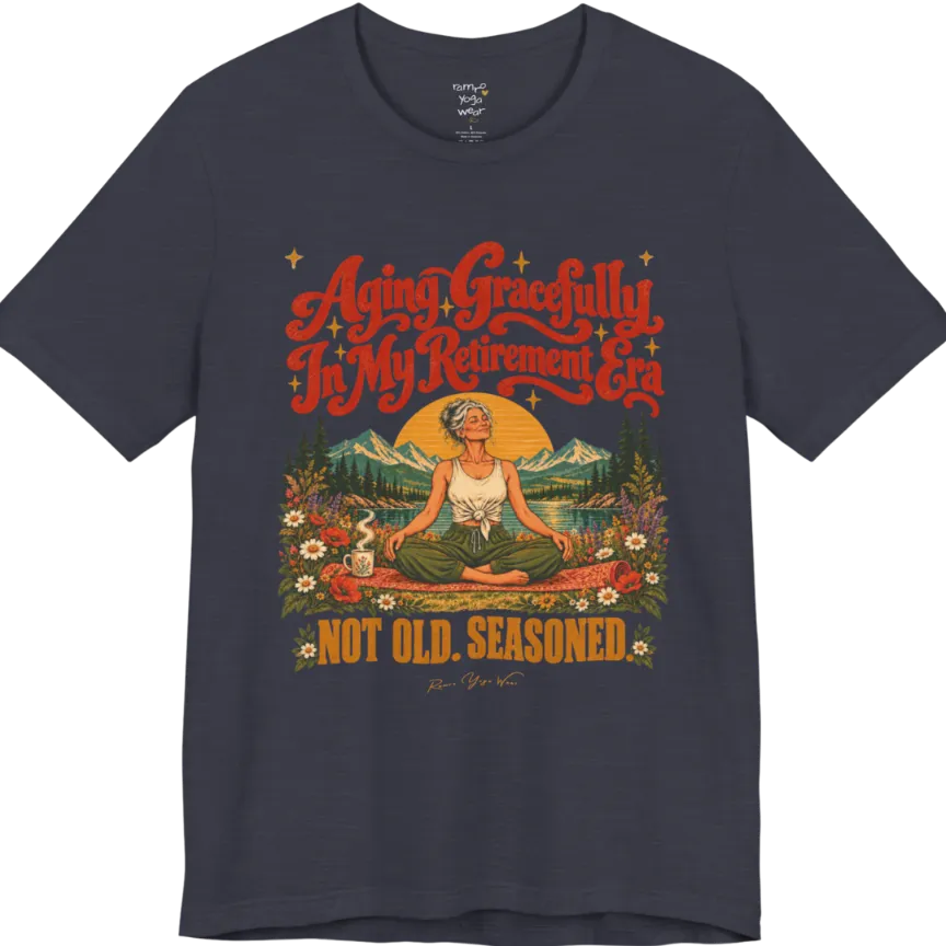 A short sleeved tee called the Seasoned T-Shirt, flat lay and a soft cotton blend shirt. Simply says, Aging Gracefully In My Retirement Era. Not Old. Seasoned. Design highlights a silver-haired figure sitting cross-legged in easy pose on a patterned mat beside a steaming mug, with a rising sun, mountains, a lake and wildflowers, and gold sparkle stars around the script lettering. Color is Heather Midnight Navy.