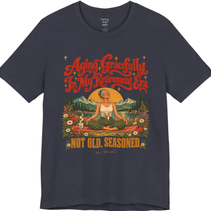 A short sleeved tee called the Seasoned T-Shirt, flat lay and a soft cotton blend shirt. Simply says, Aging Gracefully In My Retirement Era. Not Old. Seasoned. Design highlights a silver-haired figure sitting cross-legged in easy pose on a patterned mat beside a steaming mug, with a rising sun, mountains, a lake and wildflowers, and gold sparkle stars around the script lettering. Color is Heather Midnight Navy.