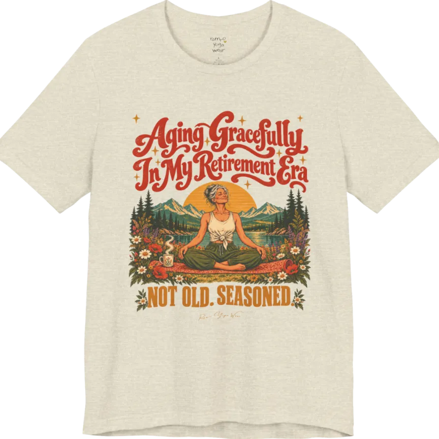 A short sleeved tee called the Seasoned T-Shirt, flat lay and a soft cotton blend shirt. Simply says, Aging Gracefully In My Retirement Era. Not Old. Seasoned. Design highlights a silver-haired figure sitting cross-legged in easy pose on a patterned mat beside a steaming mug, with a rising sun, mountains, a lake and wildflowers, and gold sparkle stars around the script lettering. Color is Heather Prism Natural.