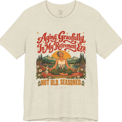 A short sleeved tee called the Seasoned T-Shirt, flat lay and a soft cotton blend shirt. Simply says, Aging Gracefully In My Retirement Era. Not Old. Seasoned. Design highlights a silver-haired figure sitting cross-legged in easy pose on a patterned mat beside a steaming mug, with a rising sun, mountains, a lake and wildflowers, and gold sparkle stars around the script lettering. Color is Heather Prism Natural.