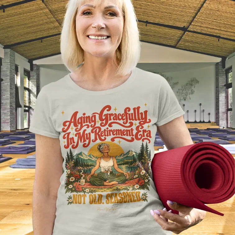 A smiling person with a blonde bob holding a rolled burgundy yoga mat, wearing a light heather Seasoned T-Shirt with a retro illustrated easy pose design, standing in a bright wooden-floored yoga studio.