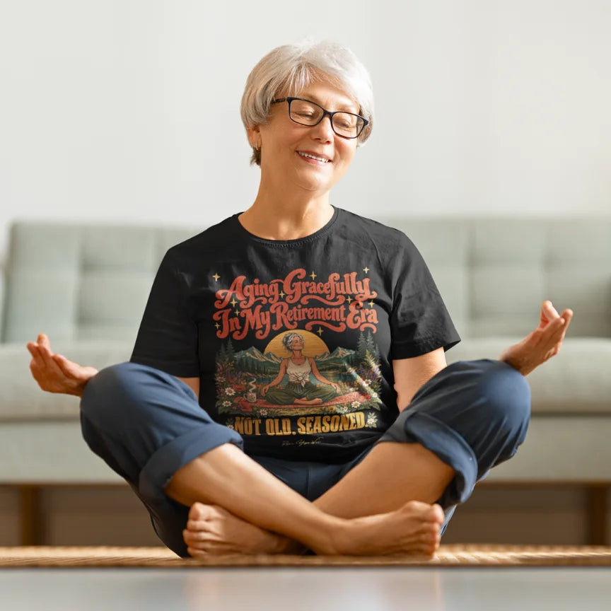 A smiling person with short silver hair and black-framed glasses, seated cross-legged in easy pose with hands resting on their knees, wearing a black Seasoned T-Shirt with a retro illustrated easy pose design, in a bright living room.