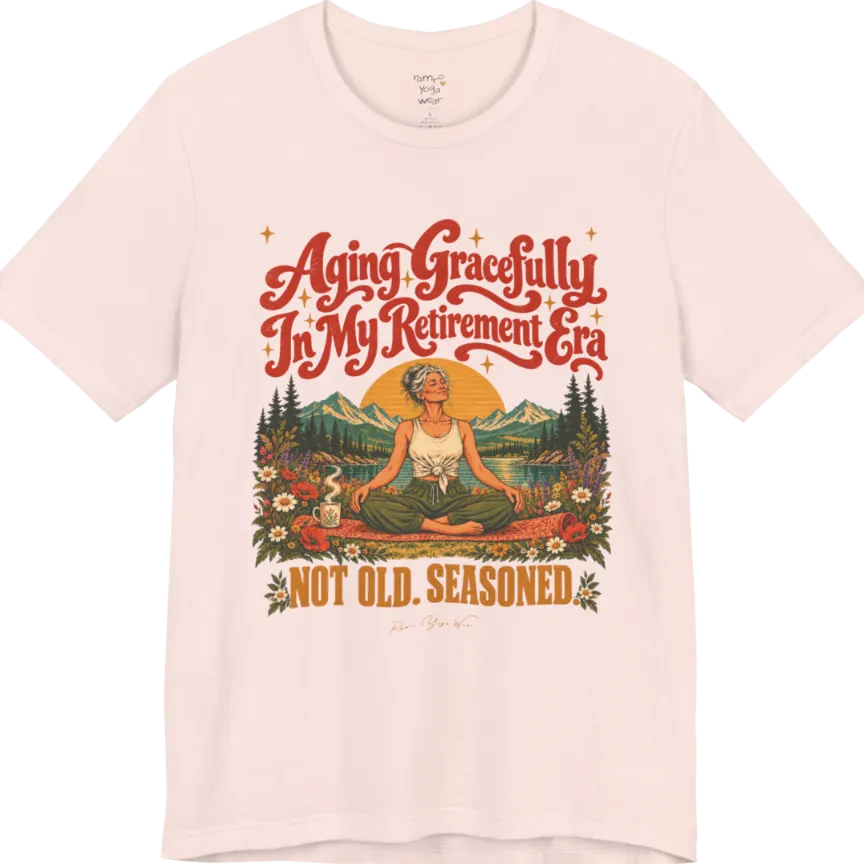 A short sleeved tee called the Seasoned T-Shirt, flat lay and a soft cotton blend shirt. Simply says, Aging Gracefully In My Retirement Era. Not Old. Seasoned. Design highlights a silver-haired figure sitting cross-legged in easy pose on a patterned mat beside a steaming mug, with a rising sun, mountains, a lake and wildflowers, and gold sparkle stars around the script lettering. Color is Soft Pink.